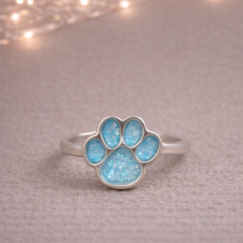 Silver ring with a blue paw print design on a textured surface with string lights in the background. a memorial ring for pets