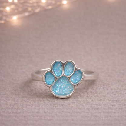 Silver ring with a blue paw print design on a textured surface with string lights in the background. a memorial ring for pets